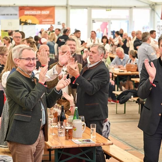Eine Gruppe von Menschen in einer festlichen Umgebung, die klatschen und lächeln, mit Tischen voller Getränke und Essen, und Banner im Hintergrund.