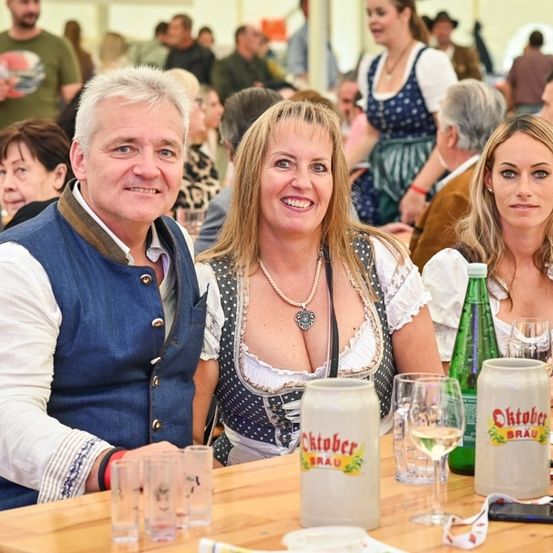 Eine Gruppe von Menschen sitzt in einem Biergarten an einem Tisch, lächelt und posiert für ein Foto. Auf dem Tisch stehen Bierkrüge, Weingläser und Flaschen. Die Frau in der Mitte trägt eine Halskette und eine gepunktete Bluse.