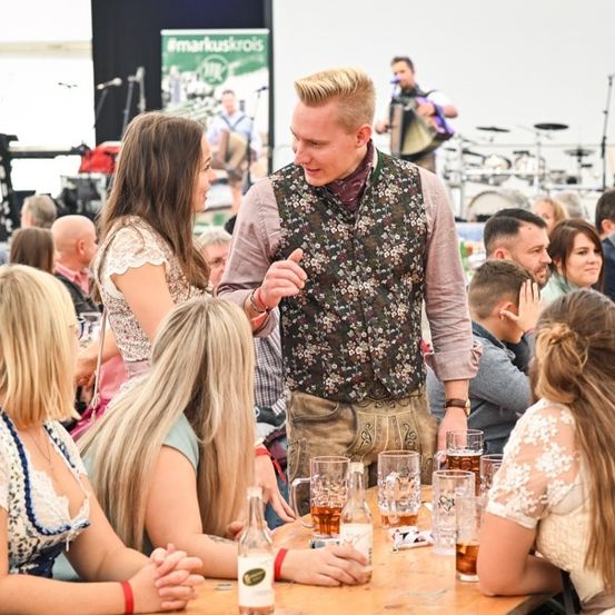 Eine Gruppe von Menschen sitzt an einem Tisch bei einer Veranstaltung im Freien, ein Mann in einer Blumenweste steht und spricht mit einer Frau. Hinter ihnen spielt eine Band auf der Bühne.