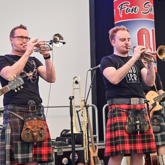 Zwei Männer in Kilts spielen Trompeten auf einer Bühne, mit einem Schlagzeuger und Gitarristen im Hintergrund.