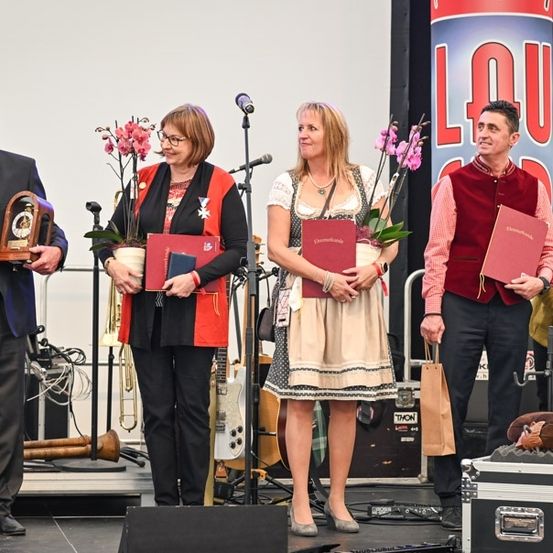 Fünf Personen stehen auf einer Bühne, halten rote Ordner und Blumen, mit einem Mikrofon und Musikinstrumenten im Hintergrund.