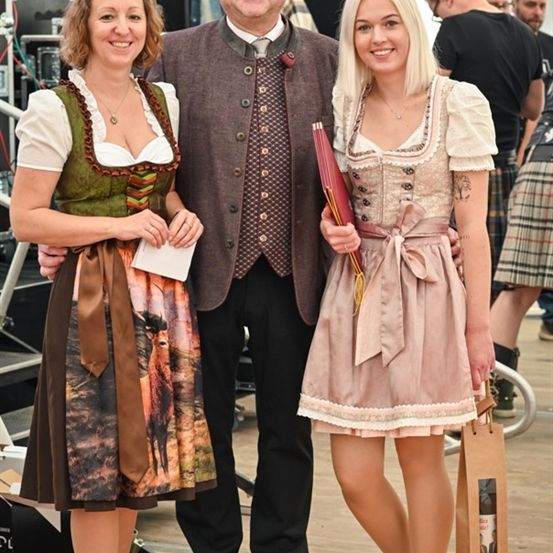 Drei Menschen stehen lächelnd bei einem Festival zusammen. Ein Mann in traditioneller Kleidung und zwei Frauen in traditionellen Kleidern.