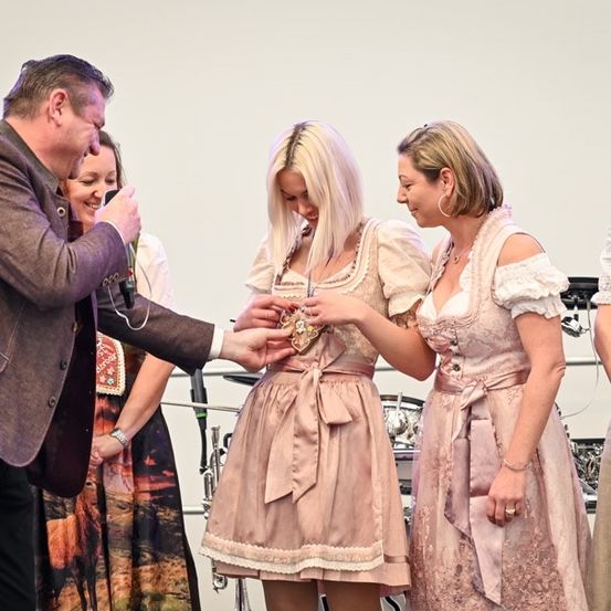 Ein Mann in einem Anzug überreicht einer Frau in einem traditionellen Kleid eine Medaille während einer Veranstaltung. Andere Frauen in traditionellen Kleidern stehen in der Nähe.