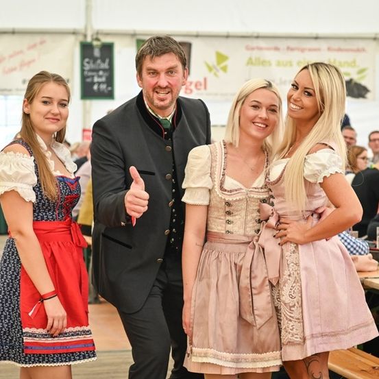 Drei Personen, zwei Frauen in traditioneller deutscher Kleidung und ein Mann in formeller Kleidung, lächeln und posieren für ein Foto bei einer Veranstaltung im Freien. Hinter ihnen sitzen Menschen an Tischen mit Getränken.