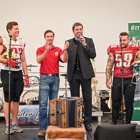 Vier Männer stehen auf einer Bühne mit Musikinstrumenten. Zwei sind Footballspieler, einer hält ein Mikrofon und einer spielt Akkordeon. Dahinter steht ein Schlagzeug und ein Banner.