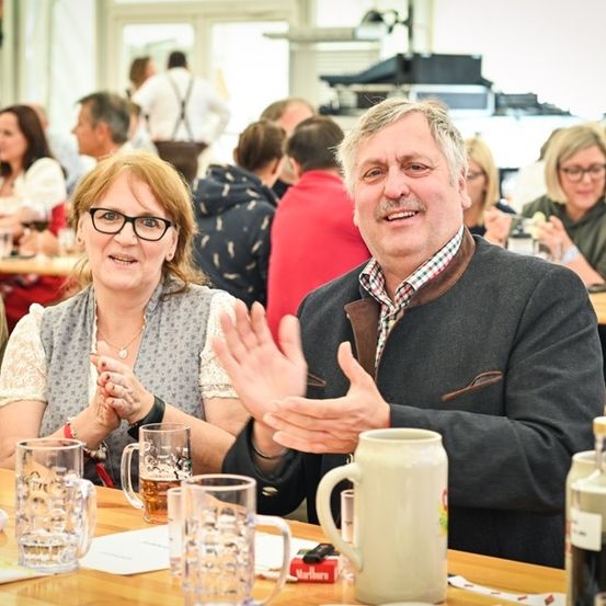 Eine Gruppe von Menschen sitzt in einem Restaurant an einem Tisch, lächelt und klatscht. Der Tisch ist mit Gläsern, Bechern und Flaschen gedeckt. Der Mann trägt eine graue Jacke und die Frau trägt eine graue Weste.