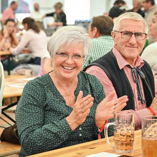 Ein älteres Paar sitzt an einem Tisch in einem Biergarten, lächelt und klatscht in die Hände. Sie sind von anderen Menschen umgeben, die ihre Getränke und Speisen genießen.