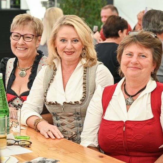 Drei Frauen in traditioneller bayerischer Kleidung sitzen an einem Tisch, lächeln und posieren für ein Foto. Sie sind von anderen Menschen umgeben, von denen einige ebenfalls traditionelle Kleidung tragen. Auf dem Tisch stehen Gläser und eine Flasche.