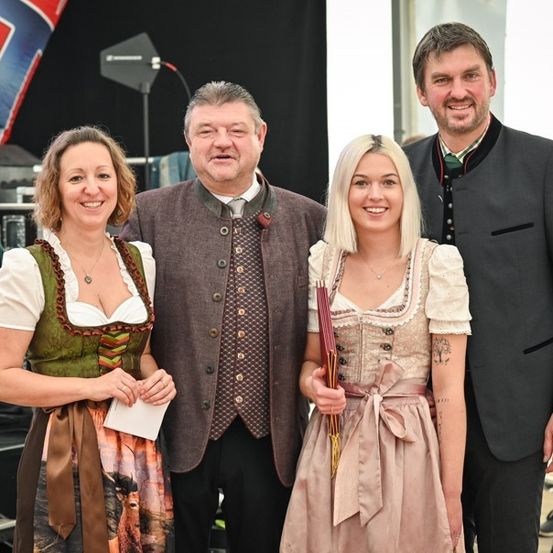 Drei Menschen lächeln und posieren für ein Foto. Die Frau links trägt ein grünes Kleid. Der Mann in der Mitte trägt eine graue Weste. Die Frau rechts hält ein Buch.