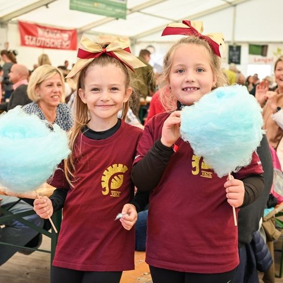 Zwei junge Mädchen, die passende weinrote T-Shirts mit gelben Schleifen auf den Köpfen tragen, halten große blaue Zuckerwatte. Sie stehen bei einer geschäftigen Outdoor-Veranstaltung, mit Menschen, die hinter ihnen sitzen und stehen.