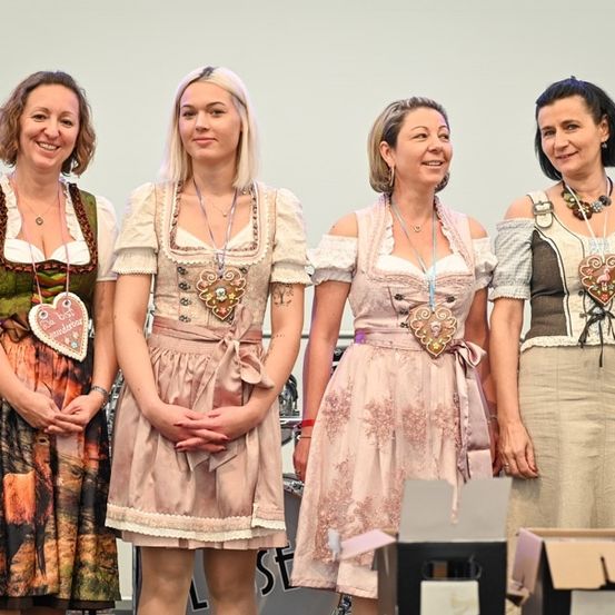 Vier Frauen in traditioneller bayerischer Kleidung stehen gemeinsam auf einer Bühne, lächeln und schauen in die Kamera. Jede Frau trägt ein anderes Kleid, eine trägt ein Herzanhänger. Im Hintergrund sind verschwommen ein Schlagzeug und Kisten zu sehen.