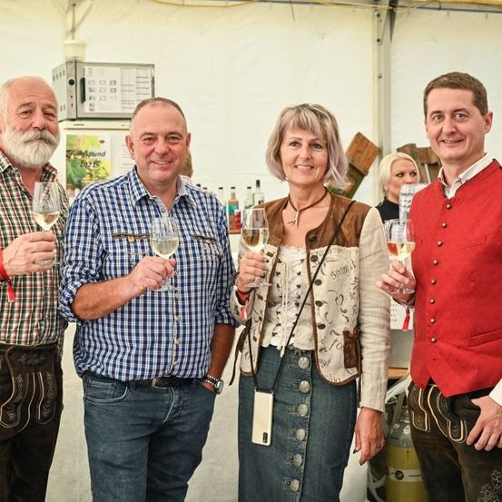 Vier Personen in traditioneller deutscher Kleidung, Weinglas in der Hand, lächeln für ein Foto. Hinter ihnen steht ein Tisch mit Flaschen und ein weißes Zelt.