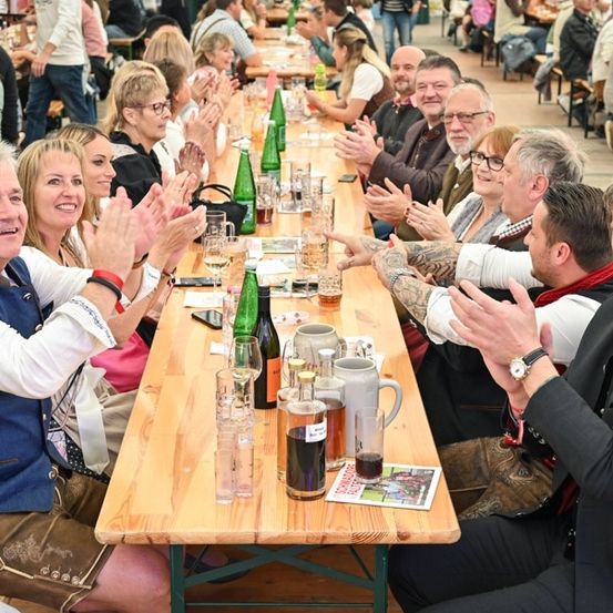 Eine Gruppe von Erwachsenen sitzt an einem langen Holztisch, klatscht und lächelt. Der Tisch ist mit Gläsern, Flaschen und einer Speisekarte gedeckt. Die Umgebung scheint ein festliches Outdoor-Event zu sein.