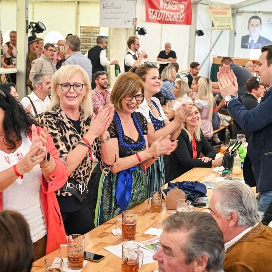 Eine Gruppe von Menschen ist auf einem Festival versammelt, klatscht und lächelt, mit Getränken und Essen auf einem Tisch vor ihnen.
