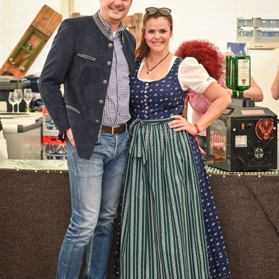 Ein Mann und eine Frau lächeln und posieren für ein Foto in einer festlichen Umgebung, mit einem Tisch und verschiedenen Gegenständen im Hintergrund.
