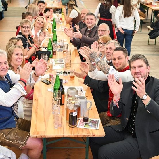 Eine Gruppe von Menschen in traditioneller bayerischer Kleidung sitzt an einem langen Tisch in einer Bierhalle, klatscht und lächelt. Der Tisch ist mit verschiedenen Getränken und Speisen gefüllt.