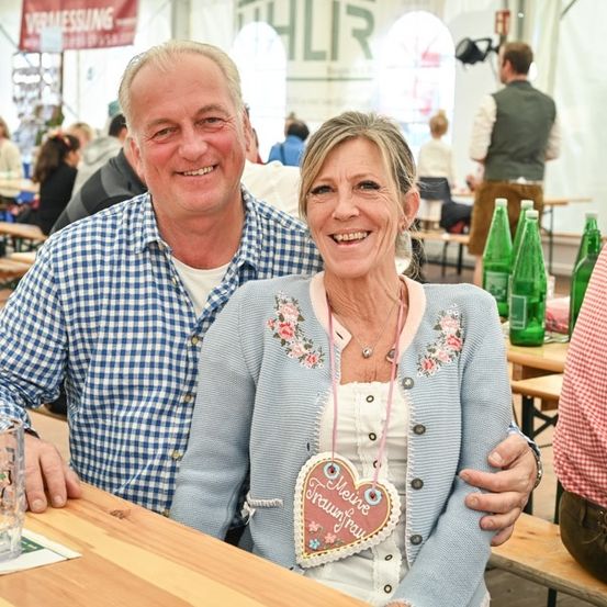 Ein Paar lächelt und posiert für ein Foto in einem Biergarten mit Holzbänken, Tischen und grünen Flaschen im Hintergrund.