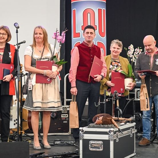 Fünf Personen stehen auf einer Bühne und halten rote Bücher und Blumen, mit einem Mikrofon, einer Aufbewahrungsbox und Musikinstrumenten im Hintergrund.