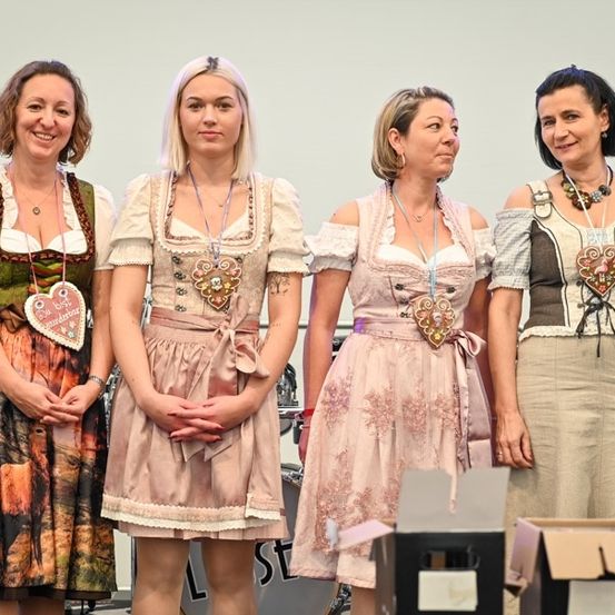 Vier Frauen in traditioneller deutscher Kleidung stehen auf einer Bühne und lächeln für ein Foto. Jede trägt eine herzförmige Plakette mit Text. Hinter ihnen sind ein Schlagzeug und Kisten zu sehen.