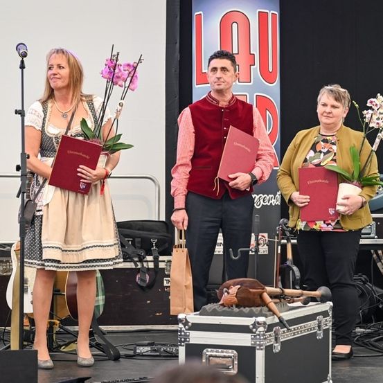 Drei Personen stehen auf einer Bühne mit Musikinstrumenten und einem Banner im Hintergrund. Die Frau links hält ein rotes Buch und Blumen, der Mann in der Mitte hält ein rotes Buch und die Frau rechts hält ein rotes Buch und Blumen.