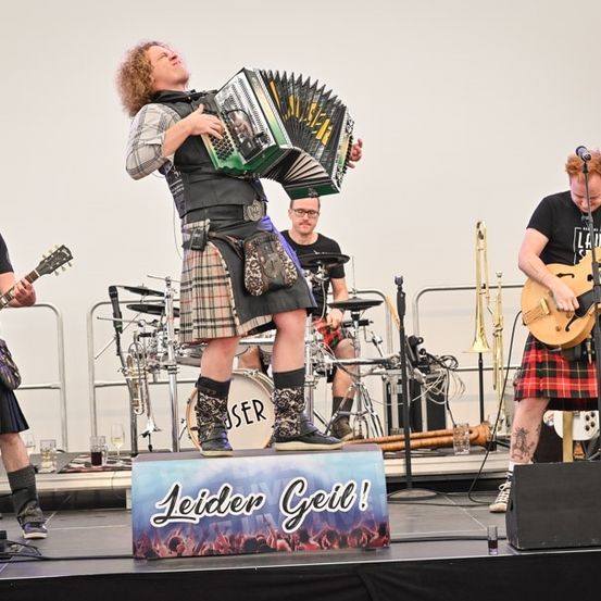 Eine Band spielt auf der Bühne mit einem Mann, der Akkordeon spielt, zwei Gitarristen und einem Schlagzeuger. Die Bühne ist mit einem Schild dekoriert, das Leider Geil! steht.