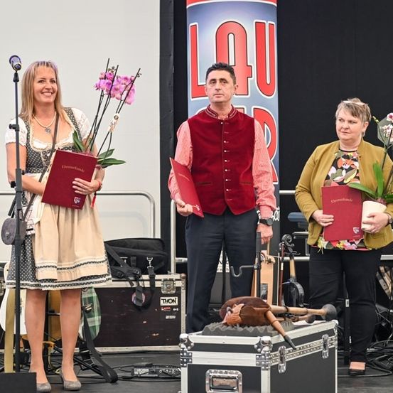 Drei Personen stehen auf einer Bühne, zwei Frauen und ein Mann, alle halten rote Bücher. Die Frau auf der linken Seite trägt ein traditionelles Kleid und hält Blumen. Der Mann in der Mitte trägt eine rote Weste. Die Frau auf der rechten Seite trägt eine gelbe Jacke. Eine Gitarre ruht auf einem Koffer vor ihnen.