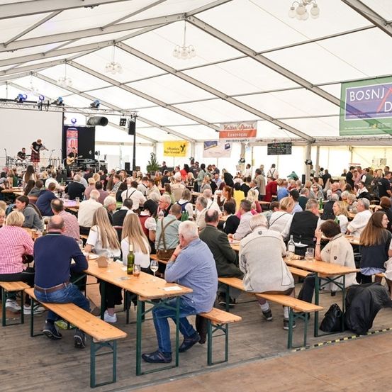 Eine große Menschenmenge sitzt unter einem Zelt an Tischen und genießt Essen und Getränke. Vorne steht eine Bühne mit Musikern, und an den Wänden hängen Banner.
