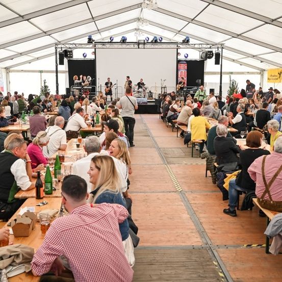 Eine große Menschenmenge sitzt in einem Zelt an Tischen und genießt Essen und Getränke, während eine Band auf der Bühne spielt. Das Zelt ist gut beleuchtet mit Deckenleuchten und Banner sind an den Wänden sichtbar.