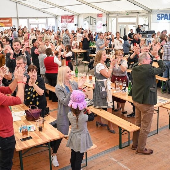 Eine große Gruppe von Menschen ist in einem Zelt versammelt, klatscht und jubelt. Einige sitzen an langen Tischen mit Getränken und Essen, während andere stehen und sich miteinander unterhalten. Das Zelt ist mit Bannern dekoriert und hat einen Holzboden.