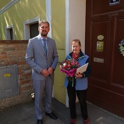 Ein Mann in einem Anzug steht neben einer Frau, die Blumen und ein Zertifikat vor einem gelben Gebäude hält.