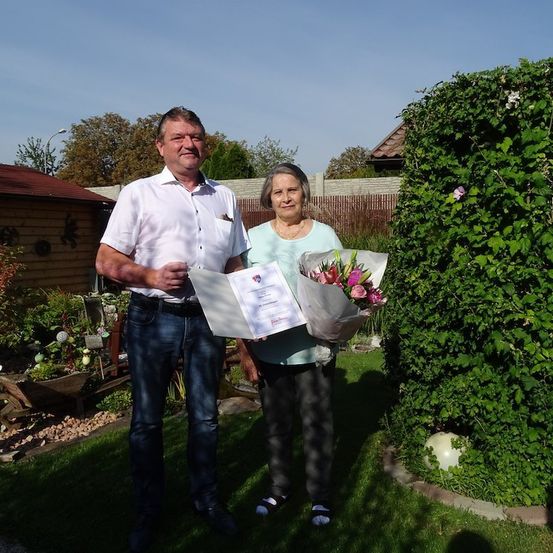 Ein Mann und eine Frau stehen in einem Garten, der Mann hält ein Zertifikat und einen Blumenstrauß, die Frau lächelt.