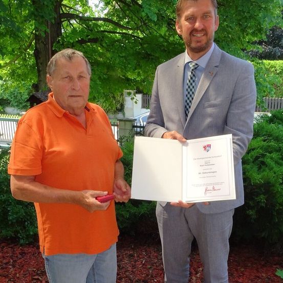 Ein Mann in einem orangefarbenen T-Shirt und ein anderer in einem grauen Anzug halten ein Zertifikat in einem Garten.