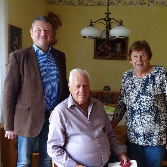 Drei Menschen stehen in einem Raum. Der Mann links trägt einen braunen Mantel, der Mann in der Mitte trägt ein lila Hemd und die Frau rechts trägt ein Blumenhemd. Hinter ihnen ist eine gelbe Wand mit einem Bilderrahmen und einer Kronleuchter.