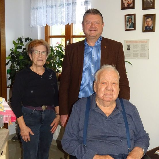 Drei Personen, zwei stehend und eine sitzend, posieren für ein Foto in einem Raum mit weißer Wand, einem Fenster mit Vorhängen und gerahmten Bildern.
