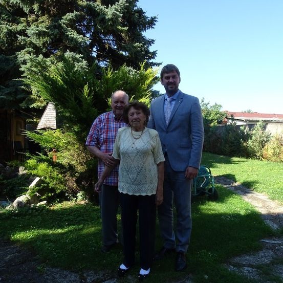 Drei Menschen stehen in einem Garten und lächeln für ein Foto. Der Mann links trägt ein kariertes Hemd, die Frau in der Mitte eine weiße Bluse und der Mann rechts einen blauen Anzug.
