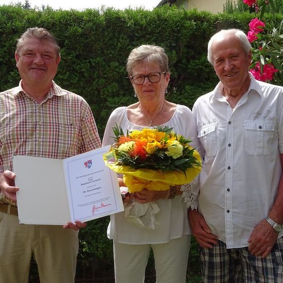Drei ältere Menschen stehen lächelnd zusammen, einer hält ein Zertifikat und einen Blumenstrauß. Die Frau in der Mitte trägt eine Brille und eine Halskette. Dahinter steht eine grüne Hecke mit rosa Blumen.
