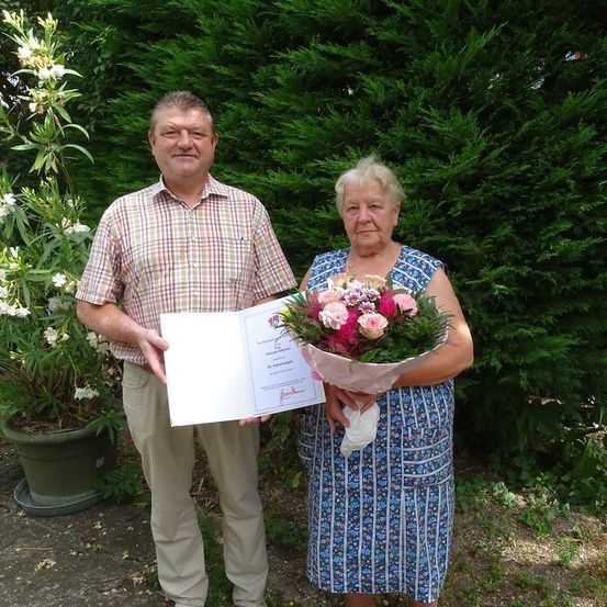 Ein älteres Paar steht in einem Garten, der Mann hält ein Zertifikat und die Frau einen Blumenstrauß. Sie lächeln und stehen nebeneinander.