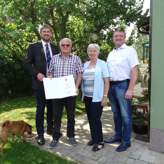 Vier Personen stehen auf einer Ziegelterrasse, eine hält ein Zertifikat. Ein Hund frisst aus einer Schüssel in der Nähe. Bäume und Pflanzen im Hintergrund.