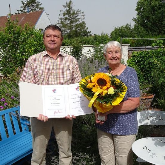 Ein Mann und eine Frau stehen in einem Garten, halten ein Zertifikat und einen Blumenstrauß. Der Mann trägt ein kariertes Hemd, und die Frau hält den Strauß mit einem Lächeln. Hinter ihnen steht eine blaue Bank und ein weißer Tisch.