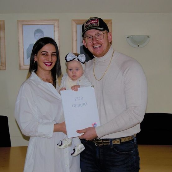 Ein Mann und eine Frau mit einem Baby halten ein Zertifikat mit der Aufschrift 'Zur Geburt'. Sie stehen vor einer Wand mit gerahmten Bildern und einem Tisch.