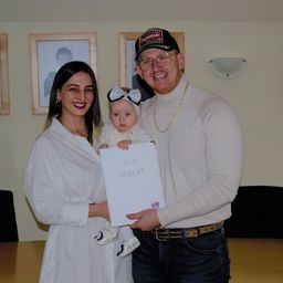 Ein Mann und eine Frau mit einem Baby halten ein Zertifikat mit der Aufschrift 'Zur Geburt'. Sie stehen vor einer Wand mit gerahmten Bildern und einem Tisch.