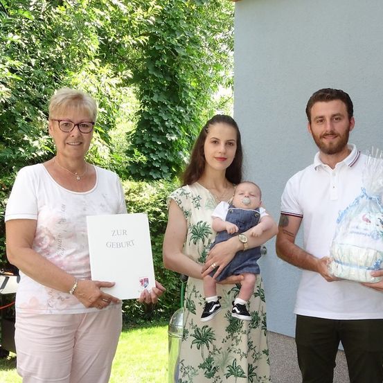 Eine Frau hält ein Zertifikat und ein Baby, eine Frau hält ein Baby und ein Mann hält ein Geschenkpaket vor einem Gebäude.