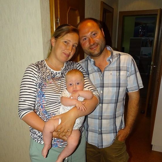 Ein lächelndes Paar mit einem Baby, der Mann in einem karierten Hemd und die Frau in einem gestreiften Shirt, stehen in einem Raum mit einer Holztür und einem Bücherregal im Hintergrund.