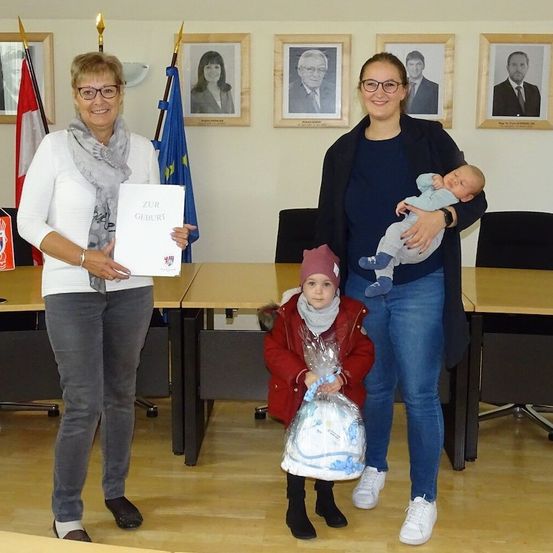 Drei Frauen und ein Baby stehen in einem Raum mit einem Tisch und Stühlen. Eine Frau hält ein Zertifikat, eine andere ein Baby und ein Kind hält ein verpacktes Geschenk. Hinter ihnen sind gerahmte Bilder und Flaggen.