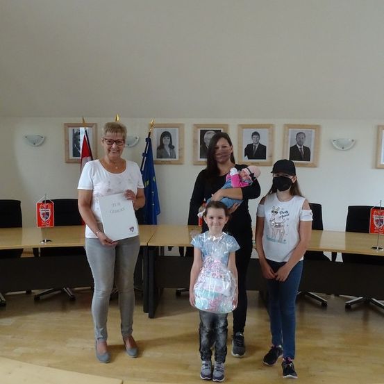 Drei Frauen und zwei Mädchen stehen in einem Raum mit einem Tisch, eine Frau hält ein Zertifikat, eine andere ein Baby und ein Mädchen ein eingewickeltes Geschenk. Hinter ihnen sind gerahmte Fotos und Flaggen.
