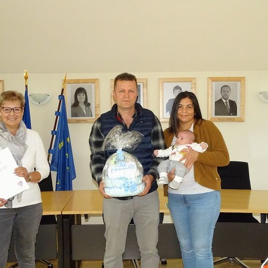 Drei Erwachsene und ein Baby stehen vor einer Wand mit gerahmten Bildern und einer blauen Flagge. Der Mann hält ein verpacktes Geschenk und die Frau hält das Baby.