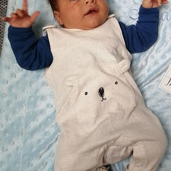 Ein Baby, das einen weißen Overall mit Bärenaufdruck trägt, liegt auf einer blauen Decke und macht mit einer Hand ein Friedenszeichen.