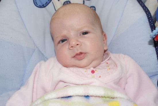 Ein Babymädchen liegt auf einer blau-weißen Decke und trägt ein rosa Outfit mit einem roten Knopf auf der Vorderseite. Sie schaut nach links mit leicht geöffnetem Mund.