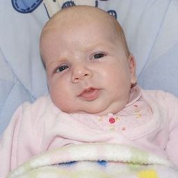 Ein Babymädchen liegt auf einer blau-weißen Decke und trägt ein rosa Outfit mit einem roten Knopf auf der Vorderseite. Sie schaut nach links mit leicht geöffnetem Mund.