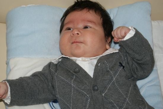 Ein Baby liegt auf einem blauen Kissen, trägt einen grauen Argyle-Pullover und ein weißes Hemd, mit einem erhobenen Arm und einem ernsten Gesichtsausdruck.